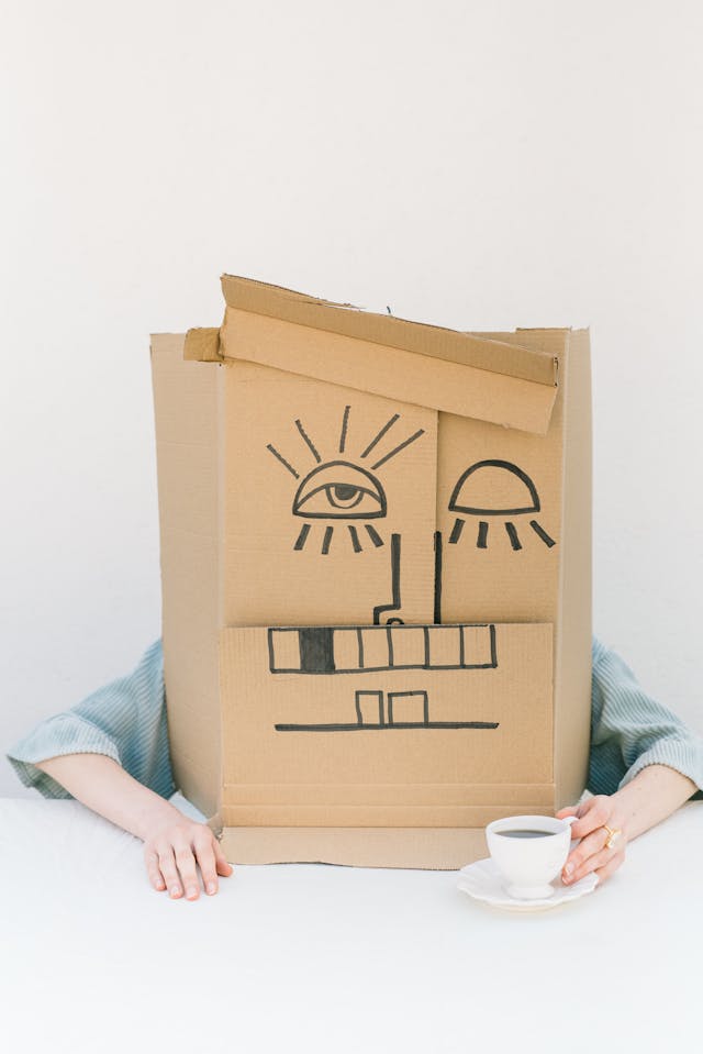 Person wearing a cardboard box with a hand-drawn face, holding a cup of coffee.