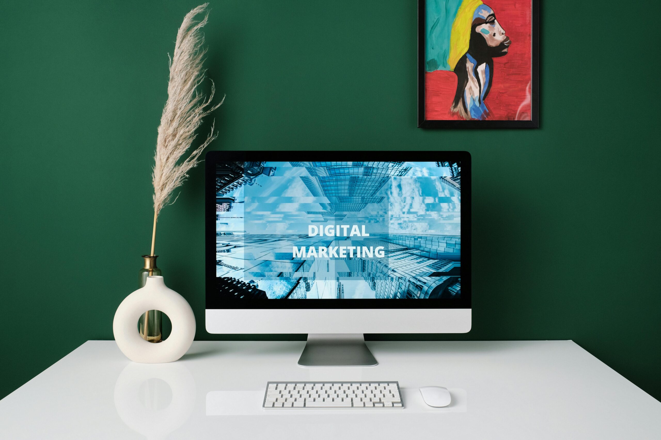 Desktop setup with a digital marketing screen, white vase, and modern art on a green wall.