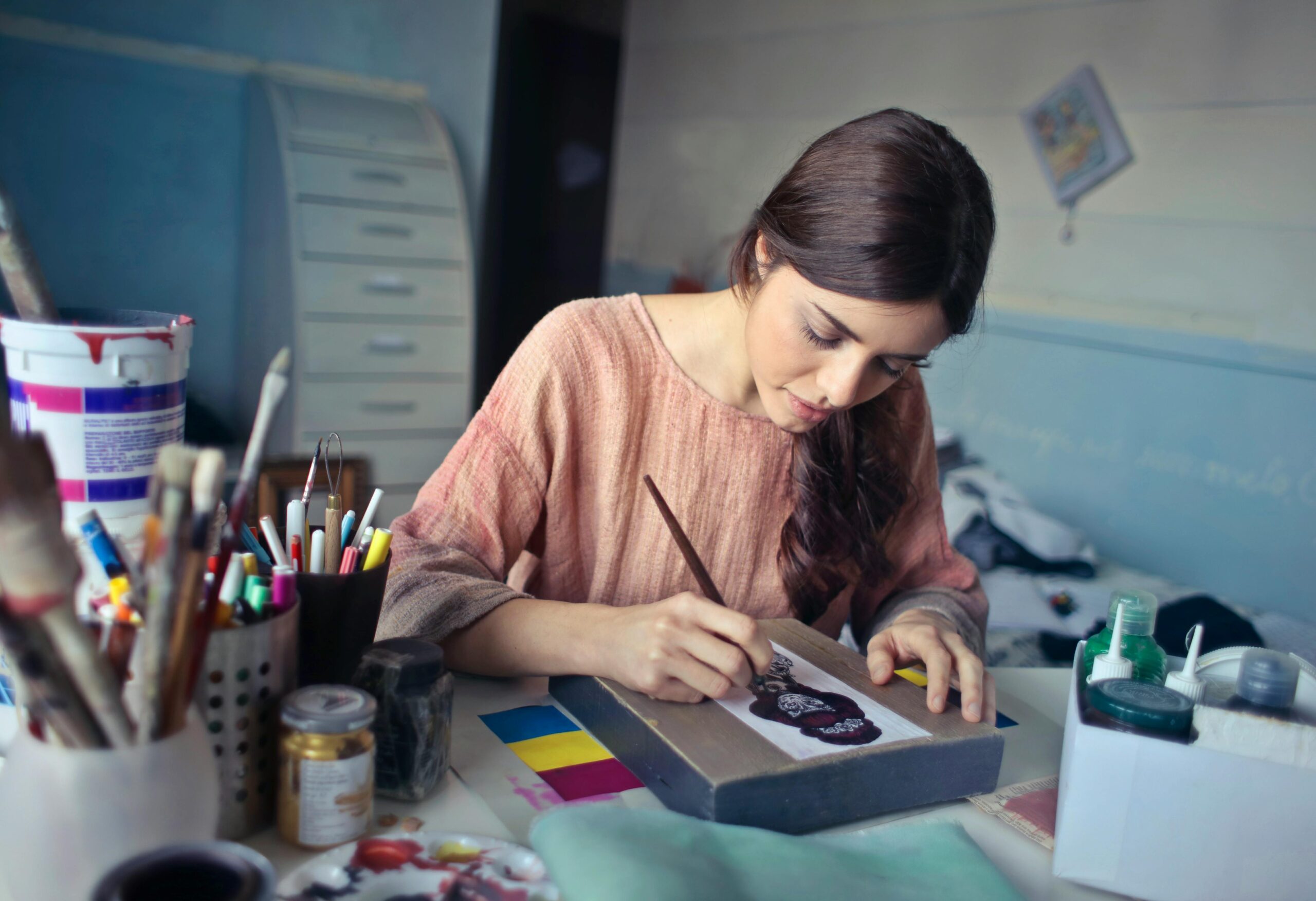 Artist focused on painting while sitting at a desk surrounded by art supplies.